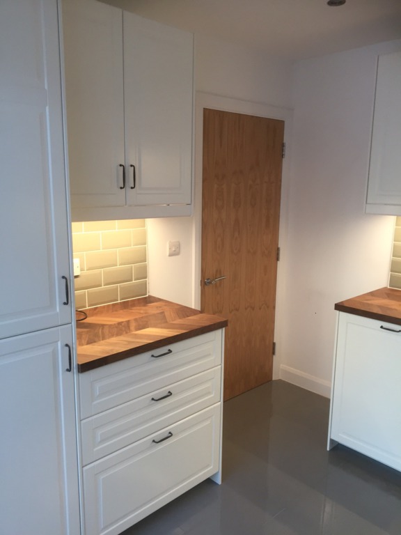 New white kitchen (shaker-style), with tiled grey floor and wooden worktops. A wooden kitchen door features in the centre of the picture